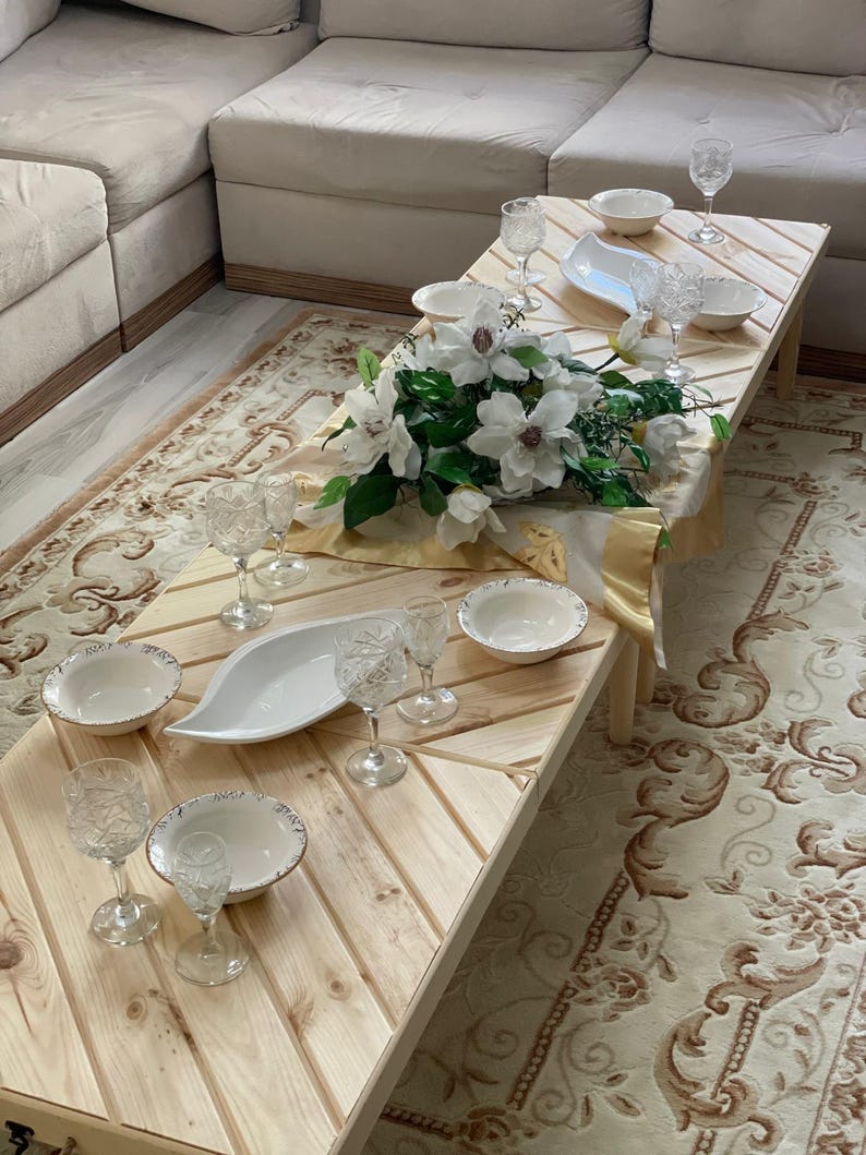 May include: A wooden table with a chevron pattern, set for a meal with white bowls, plates, and glasses. A bouquet of white flowers sits in the center of the table.