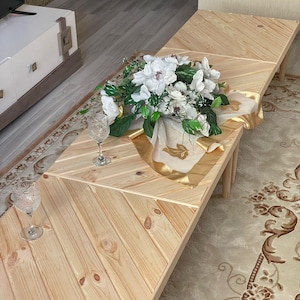 May include: A wooden coffee table with a chevron pattern. The table is made of light wood and has a natural finish. The table is decorated with a bouquet of white flowers and a gold-colored fabric.