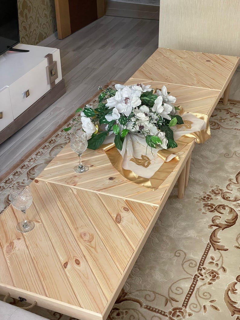 May include: A wooden coffee table with a chevron pattern. The table is made of light wood and has a natural finish. The table is decorated with a bouquet of white flowers and a gold-colored fabric.