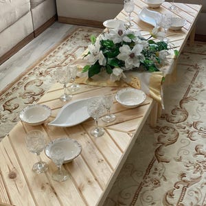 May include: A wooden table with a light wood finish and a white tablecloth. There are white bowls, clear glasses, and a white serving dish on the table. A bouquet of white flowers is in the center of the table.
