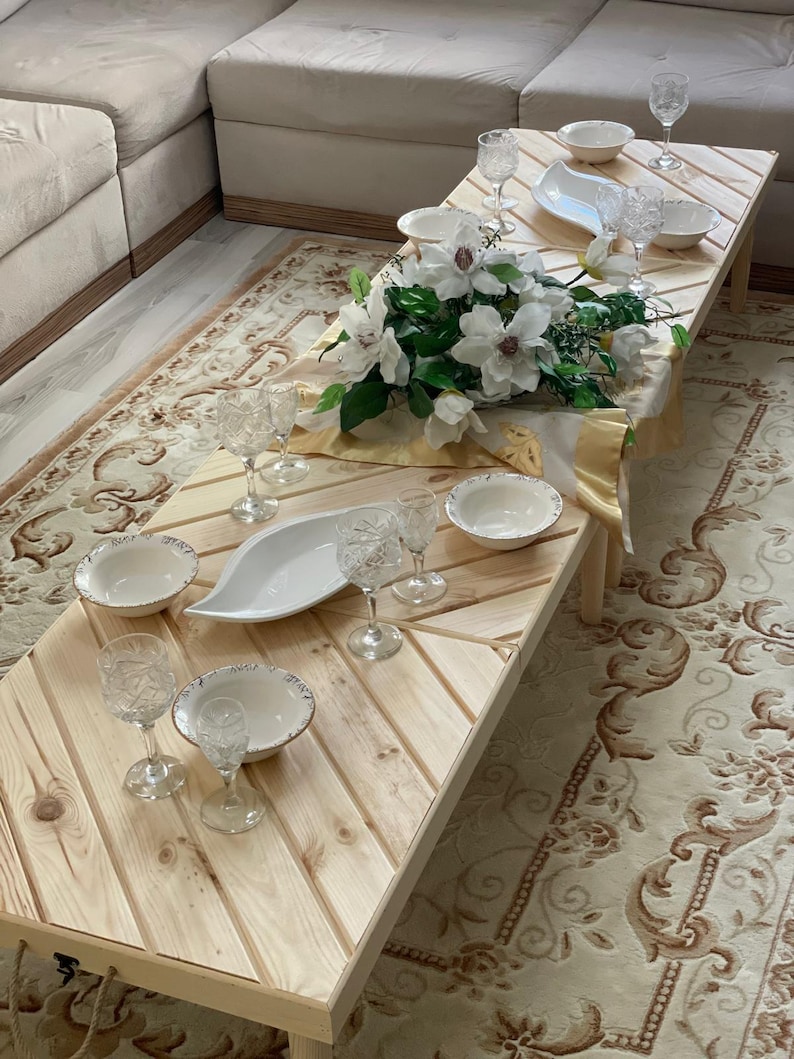 May include: A wooden table with a light wood finish and a white tablecloth. There are white bowls, clear glasses, and a white serving dish on the table. A bouquet of white flowers is in the center of the table.