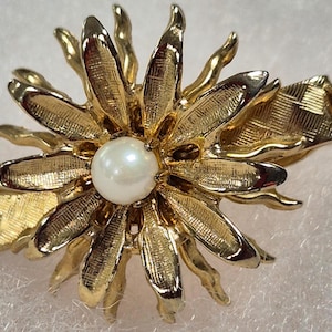 May include: A gold-toned flower-shaped brooch with a pearl center. The brooch has detailed petal designs and a textured finish. The clasp is visible on the side. This vintage-style jewelry piece is suitable for various occasions.