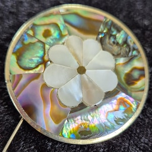 May include: A round, silver-toned brooch with a white, flower-shaped inlay. The flower is surrounded by iridescent, abalone shell pieces.