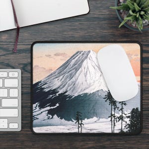 May include: A black mousepad with a Japanese-style illustration of Mount Fuji, a snow-capped mountain, with a pink and orange sunset sky in the background.  There are small trees in the foreground.