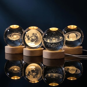 Peut inclure: Quatre sphères en verre transparent, chacune présentant un design céleste gravé au laser en 3D détaillé, incluant une galaxie, la lune, le système solaire et Saturne. Chaque sphère est éclairée par le bas par une base en bois clair et ronde.