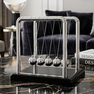 May include: A Newton's cradle featuring five polished silver-colored metal spheres suspended by thin wires from a rectangular chrome frame. The entire structure is mounted on a solid, smooth black rectangular base with a reflective finish.