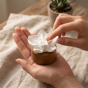Peut inclure: Un ornement floral en céramique blanche avec des pétales lisses et superposés fixés sur une base cylindrique en bois. L'objet est compact et présente une finition en grain de bois naturel sur les côtés de la base, contrastant avec la surface blanche mate.