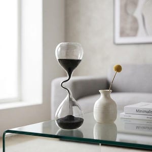 May include: A clear glass hourglass with a unique twisted neck design containing fine black sand. The hourglass is positioned next to a small, off-white ceramic vase holding a single yellow flower and a stack of white books on a glass table.