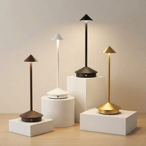 May include: Four portable table lamps with slender stems and conical shades. The lamps are finished in matte black, white, dark brown, and metallic gold. Each lamp features a circular base with a small power button and emits a soft downward light.