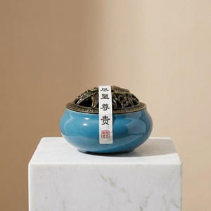 May include: A round, teal ceramic incense burner with a dark bronze-toned metal lid featuring intricate cut-out patterns. A vertical white paper strip with black Chinese characters and a small red seal is affixed to the center of the lid and bowl.