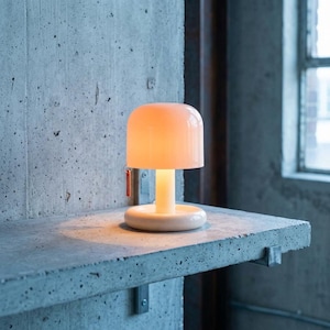Puede incluir: Una lámpara de mesa portátil con una base cilíndrica lisa de color blanco roto y una pantalla redondeada y translúcida en forma de cúpula que emite un brillo cálido y suave. Una pequeña etiqueta rectangular naranja cuelga del lateral.