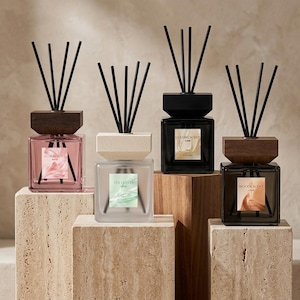 Peut inclure: Quatre diffuseurs de parfum en verre carré avec des bâtonnets noirs et des bouchons en bois. Les flacons sont roses, blancs givrés, noirs et gris foncé, avec des étiquettes Floral Note, Tea Leaves, Gulong Scent et Woode Scent.