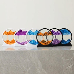 May include: Six circular sand art displays arranged in a row. Three frames are white and three are black. Each contains liquid and colored sand forming mountain-like landscapes in shades of orange, purple, and blue, resting on stable bases.