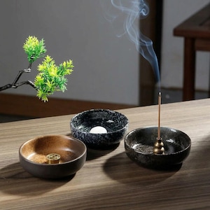 Peut inclure: Trois petits bols en céramique servant de porte-encens. Un bol est marron avec un insert en laiton, un est noir avec des taches blanches et une sphère blanche, et un est gris foncé avec un porte-bâtonnet en laiton dégageant de la fumée.