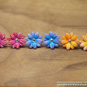 May include: Five pairs of colorful flower-shaped earrings with a rhinestone center. The earrings are pink, blue, and orange.