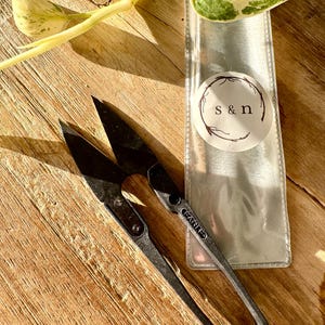 May include: Black thread snips with a curved handle and sharp blades, next to a clear plastic sleeve with a circular label that reads "s & n". The snips are on a wooden surface, with a green and yellow plant in the background.