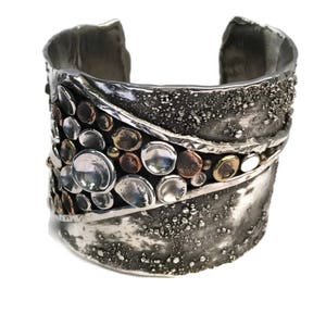 May include: A silver cuff bracelet with a textured surface and a diagonal band of silver, copper, and gold circles.