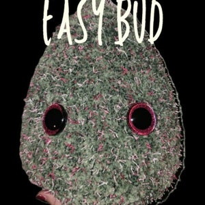 Pode incluir: Um amigurumi de flor de cannabis de crochê verde e rosa com olhos pretos e pálpebras vermelhas. O amigurumi é etiquetado como "Jumbo Easy Bud" e "Cannabis Flower Amigurumi".