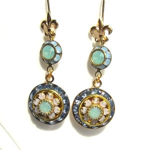 May include: A pair of gold dangle earrings with blue and white crystal accents. The earrings feature a fleur-de-lis design at the top and a round, multi-faceted crystal in the center.