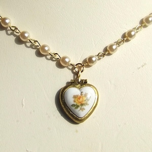May include: A gold chain necklace with a heart-shaped locket pendant. The locket is white with a floral design and a yellow rose.