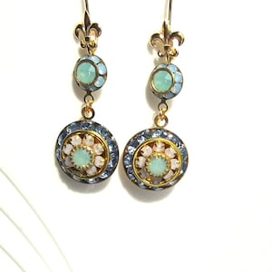 May include: A pair of gold dangle earrings with blue and white crystal accents. The earrings feature a fleur-de-lis design at the top and a round, multi-faceted crystal in the center.