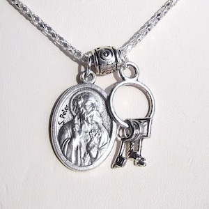 May include: Silver chain necklace with a silver oval pendant featuring a depiction of Saint Peter. The pendant is attached to a silver ring with two silver keys.