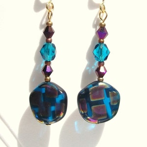 May include: A pair of dangle earrings with gold-colored hooks. Each earring features a large, square-shaped, iridescent blue bead with a grid pattern, accented by teal and purple crystal beads.