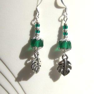 May include: A pair of dangle earrings featuring faceted emerald green beads, small silver beads, and silver-colored leaf charms. The earrings have silver-colored ear wires and a tropical theme.