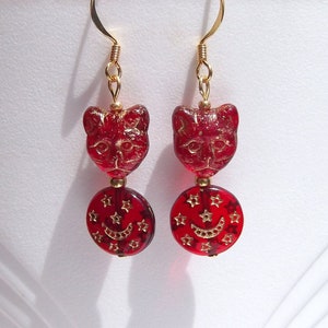 May include: A pair of red cat-shaped earrings with gold-colored accents. The earrings feature a cat face at the top and a round charm with stars and a crescent moon. The earrings are suspended from gold-colored ear wires.