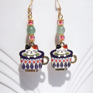 May include: A pair of gold-toned dangle earrings. Each earring features a cat in a teacup charm with a blue and white pattern, accented with red and green beads. The cat has black, white, and orange fur. The earrings have a whimsical, playful design.