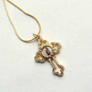 May include: A gold-colored cross pendant necklace. The cross has an ornate design with a small image at the center. The chain is a thin, gold-colored snake chain. The pendant is approximately 1.5 inches in length.