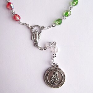 May include: A silver rosary with green, red, and clear glass beads. The rosary has a silver charm with a depiction of the Virgin Mary and a silver medal with the text "Saint Dymphna Patroness of Stress Anxiety Mental Health".