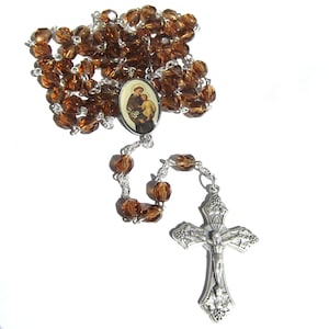 May include: A religious rosary featuring faceted amber-colored crystal beads linked by silver-toned metal chain. The centerpiece is an oval medal depicting a figure holding a child, and the crucifix is a silver-toned metal cross with a figure of Jesus.