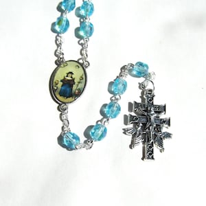 May include: A rosary featuring translucent light blue faceted beads connected by a silver-toned metal chain. It includes an oval-shaped centerpiece depicting a figure in a blue robe and a silver-toned crucifix with an engraved crossbar and angels.