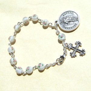 May include: A silver-toned chain bracelet featuring white beads with small pink and green floral patterns. The bracelet includes a round metal charm embossed with the text RAPHAEL ARCHANGEL and a decorative silver-toned cross pendant attached to the chain.