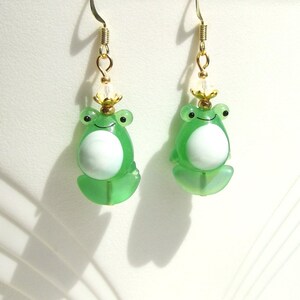 May include: A pair of whimsical frog earrings. Each earring features a translucent green frog with a white belly, black eyes, and a small gold crown. The earrings hang from gold-colored fish hooks.
