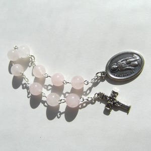 May include: A rosary featuring pale pink rose quartz beads strung on a silver chain. The rosary includes a silver cross and a silver oval pendant with an image of St. Peregrine.