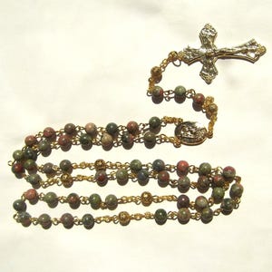 May include: A rosary with multi-colored stone beads and gold-colored chain links. The rosary features a gold-colored crucifix and a circular centerpiece. The beads are a mix of green, red, and gray.