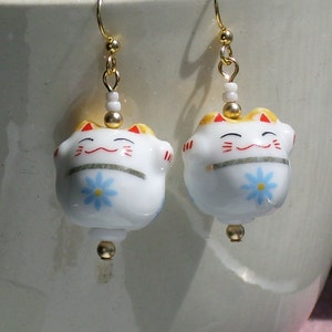 May include: A pair of gold earrings with white porcelain cat charms. The cats have a smiling face, red cheeks, and a blue flower on their belly.