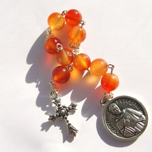 May include: A carnelian stone rosary with a silver cross and a silver charm featuring a depiction of Saint Peregrine. The charm has the text "Saint Peregrine" inscribed on it.