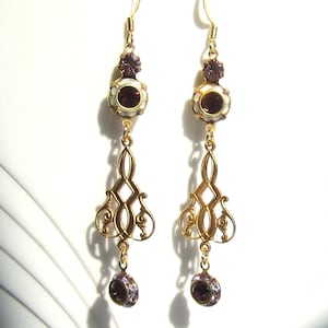 May include: Gold dangle earrings with a white and purple enamel design. The earrings feature a small round purple stone at the bottom and a larger oval purple stone at the top.