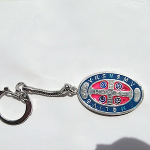 May include: A silver key chain with a blue and pink oval charm. The charm has a cross with the letters "V.R.S.N.S.M.V." around the edge and "PAX" and "M.G.L.I.V.B." on either side of the cross.