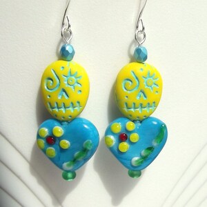 May include: A pair of handmade earrings featuring a Day of the Dead skull design. The earrings have yellow skull-shaped beads with blue accents, paired with blue heart-shaped beads adorned with flowers. Silver-tone ear wires complete the design.
