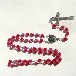 May include: A rosary with faceted red beads and a silver-toned metal cross. The beads are strung on a silver chain, and the cross has decorative accents. The rosary also features a heart-shaped centerpiece.