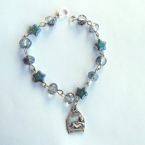 May include: A silver-toned bracelet featuring iridescent blue star-shaped and clear round beads. A small pendant with an image of a baby and the words "GLORIA DEO" hangs from the bracelet. The bracelet has a lobster clasp closure.