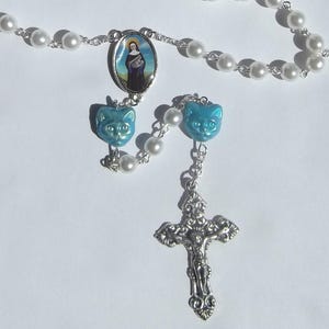 May include: A rosary featuring white pearl-like beads on a silver-toned chain, accented with two light blue cat-shaped beads. The centerpiece is an oval medal depicting a figure in a dark habit, and the crucifix is an ornate silver-toned metal.