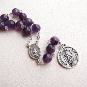 May include: Amethyst rosary with silver beads and two silver medallions. One medallion features the image of the Virgin Mary and the other features the Archangel Raphael.