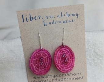 May include: A pair of handmade, hot pink embroidered earrings with silver-colored hooks. The earrings are circular with a detailed, concentric design. They are displayed on a brown card with the text "Fiber: art, alchemy, & adornment" and a website address.