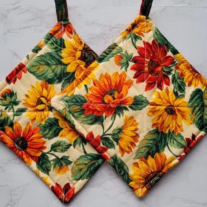 May include: Two quilted pot holders with a sunflower print. The pot holders are cream colored with red, yellow, and green sunflowers.