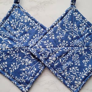 May include: Two blue quilted pot holders with a white floral pattern. The pot holders have a loop for hanging.